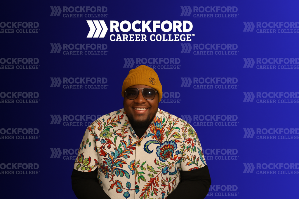 Keiland Lumpkins Sr. smiling in front of a blue Rockford Career College backdrop, wearing glasses, a yellow knit hat, and a floral shirt.