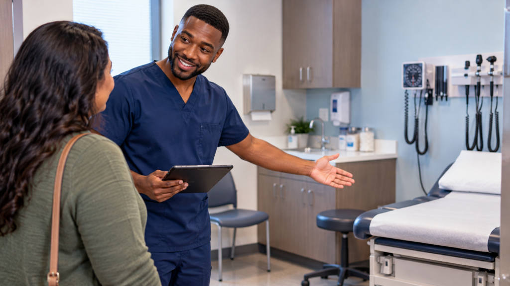 Medical assistant in navy-blue scrubs welcoming a patient into a healthcare examination room.