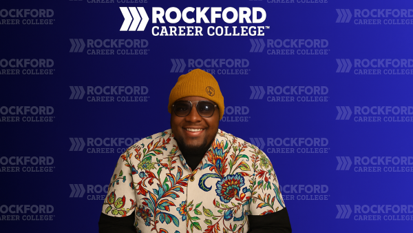 Keiland Lumpkins Sr. smiling in front of a blue Rockford Career College backdrop, wearing glasses, a yellow knit hat, and a floral shirt.